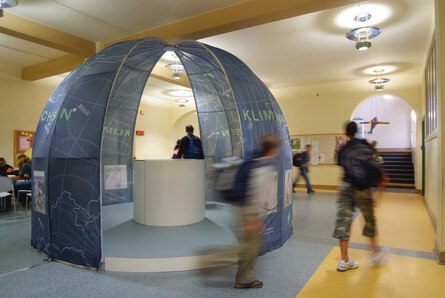 Ausstellung Klimapavillon: Foto des blauen Pavillons in einem Schulhaus, ringsherum laufen Schulkinder vorbei